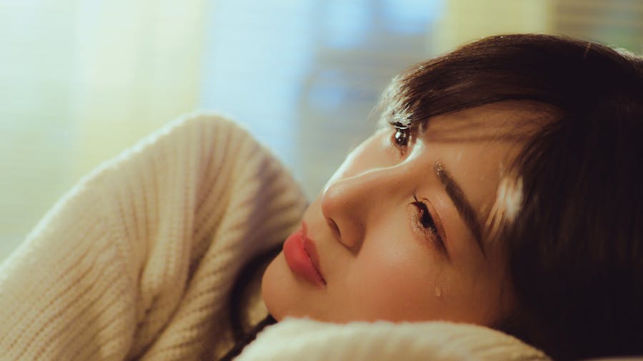 Close-up of a ladyboy lying down in a white sweater, looking upward with a tear rolling down her cheek in soft lighting.