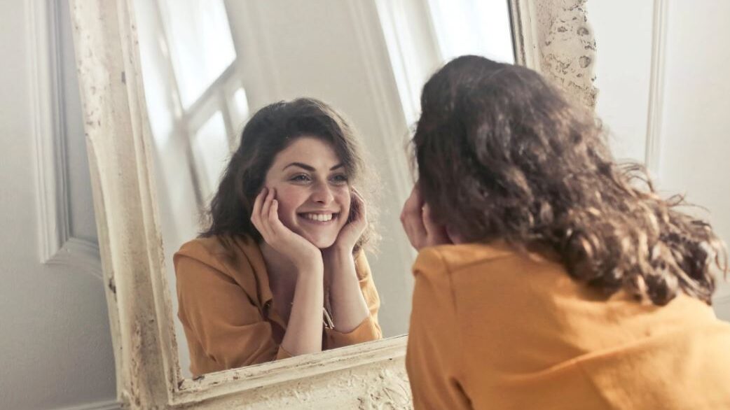 Ladyboy in an orange blouse smiling at her reflection in a vintage mirror.