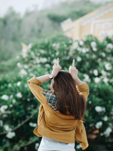 Rear view of a woman in a yellow jacket and white top making rebellious gestures in a garden.