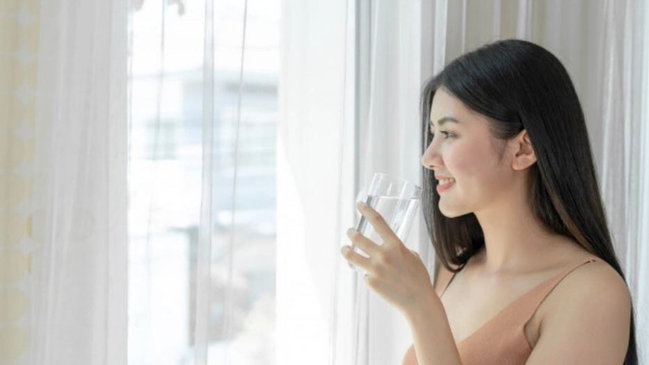 Woman drinking water from a glass by a window with white curtains.