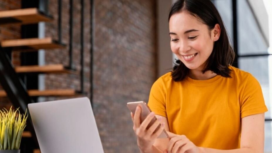 Woman smiling while looking at her phone, seated next to a laptop in an office setting.