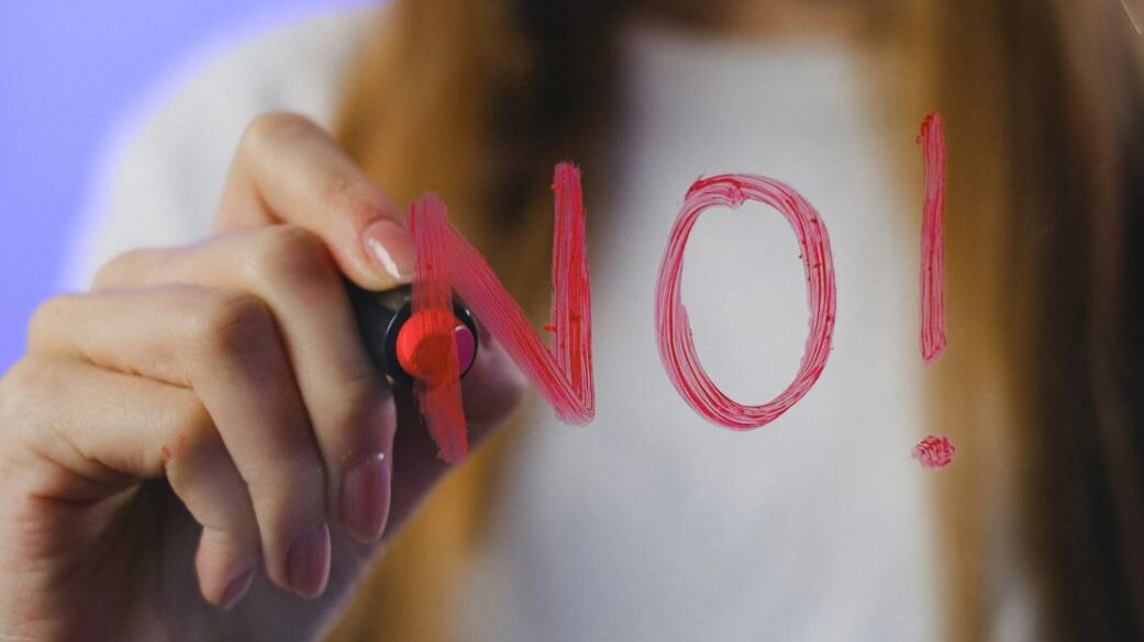 Person writing 'NO' in red marker on glass, viewed from behind the glass.