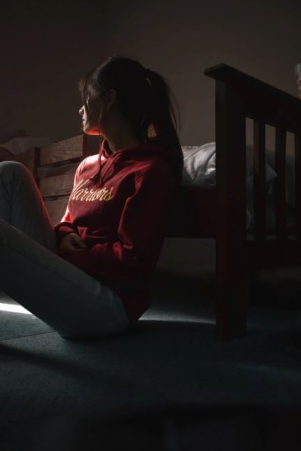 Woman sitting on the floor in a dimly lit room, looking out a window.