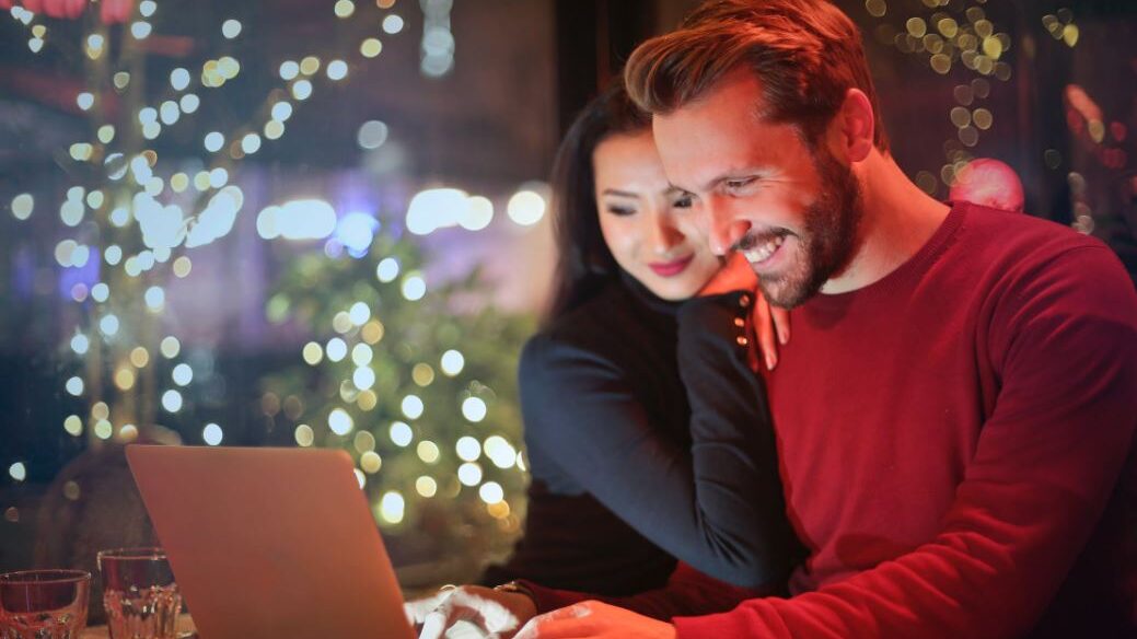 Couple smiling while looking at a laptop screen together in a cozy, festive setting.