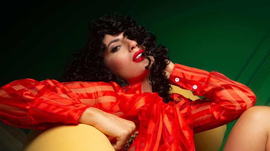 Woman with curly hair and red lipstick wearing a red blouse, posing confidently.