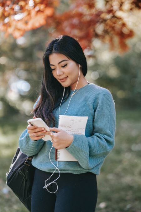 Woman holding a phone and notebook while wearing earphones.