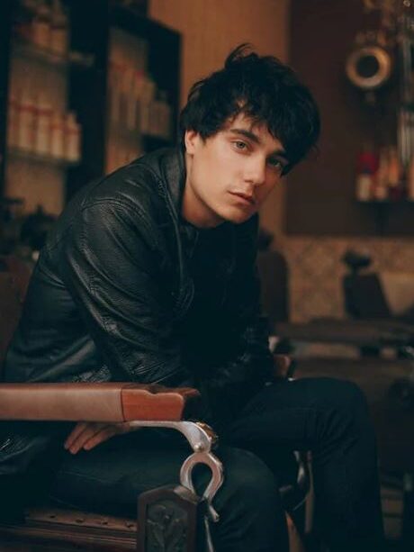 Young man sitting in a leather chair, wearing a black jacket, looking at the camera.