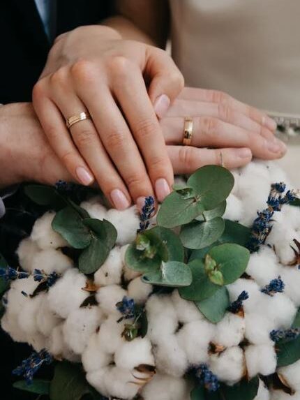 Close-up of two hands wearing wedding rings placed on top of a cotton flower bouquet.