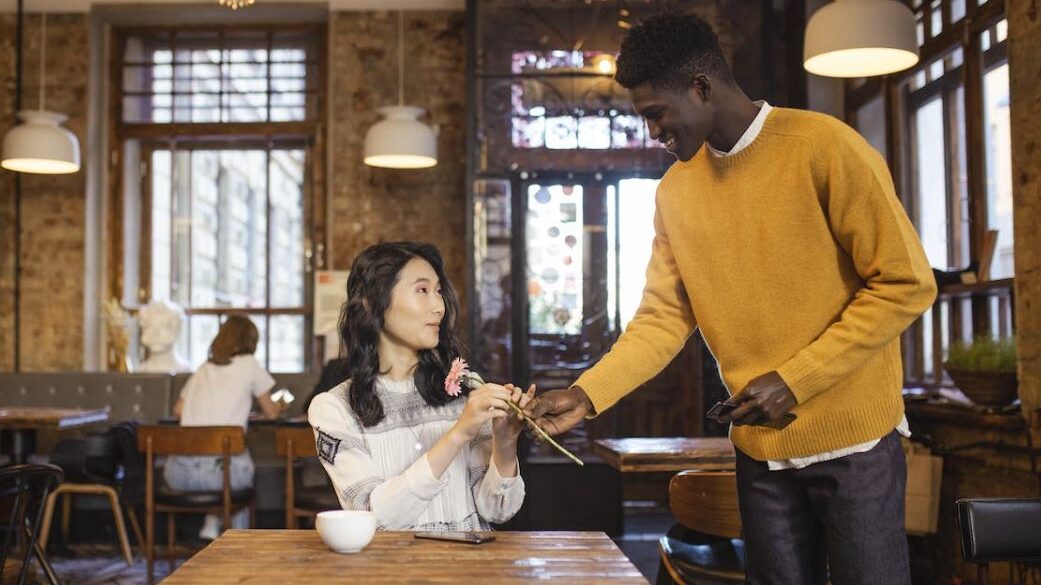 Woman accepting a flower from a man in a café, both sharing a warm interaction.