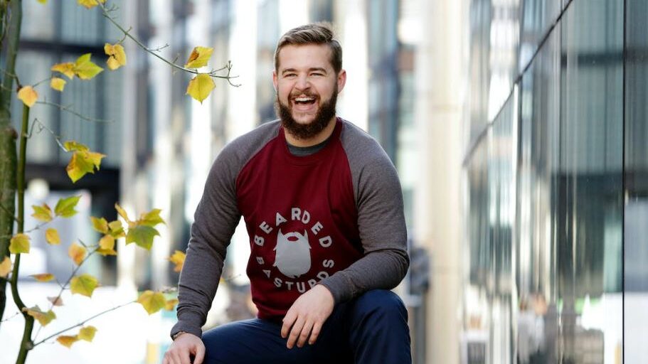 A bearded man sitting on a bench outdoors, smiling and wearing a burgundy sweatshirt.