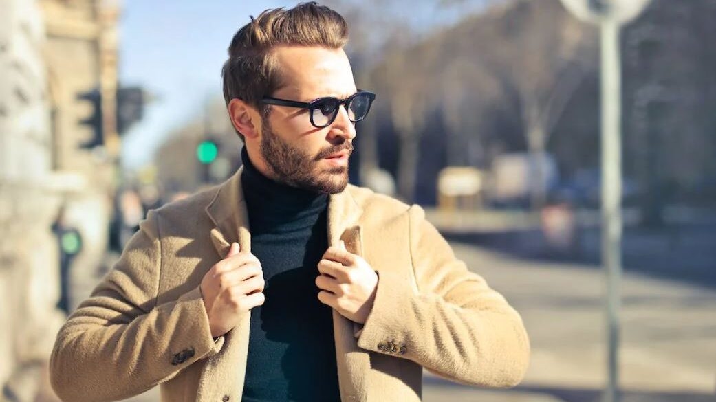 Man in a tan coat and sunglasses, standing in the street on a bright day.