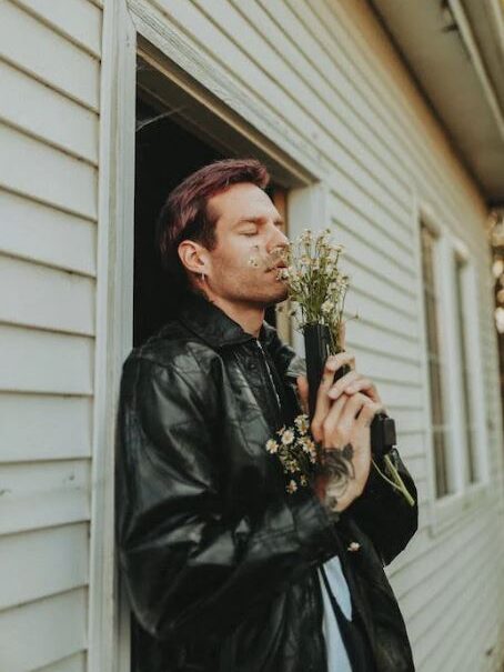 A man standing at a window, holding a bouquet of flowers and gently smelling them.