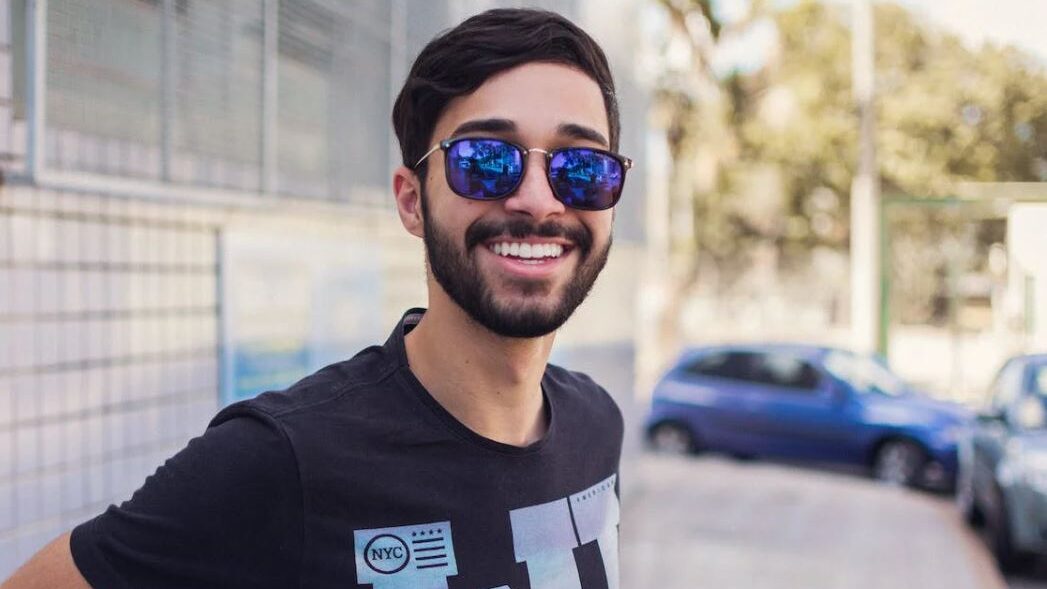 Man wearing blue mirrored sunglasses, smiling confidently in an outdoor setting.