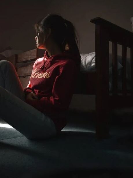 Woman sitting on the floor near a bed, partially illuminated by light coming through a window.