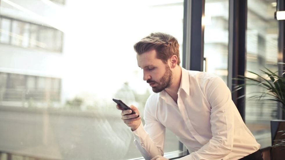 A man in a white shirt standing by a large window, looking at his phone in a focused manner.