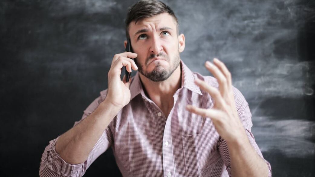 Person holding a phone to their ear, making an exasperated face and raising one hand.