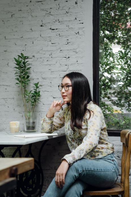 A woman wearing a floral blouse and glasses, thinking deeply as she sits near a window with plants nearby.