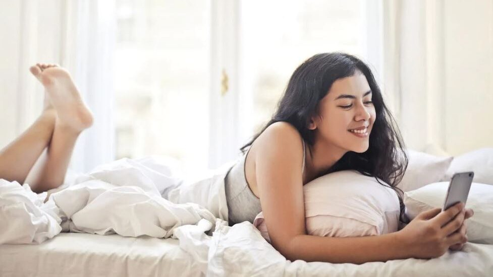 A young woman lying on her stomach on a bed, smiling as she reads something on her phone.