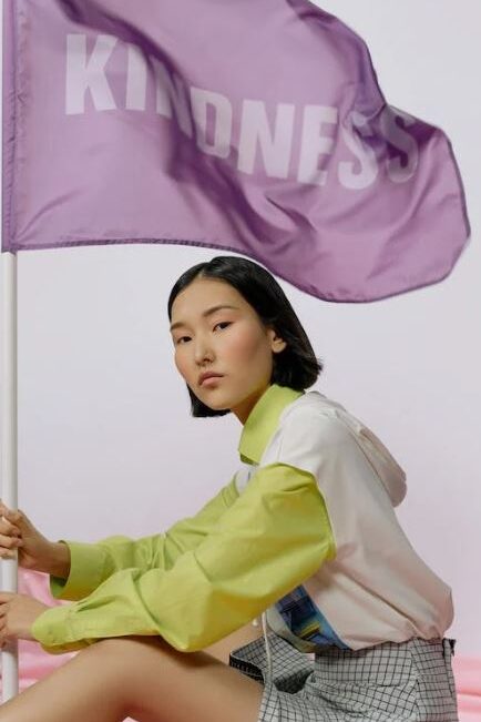 A woman holding a purple flag that says "Kindness," dressed in a trendy outfit and striking a confident pose.