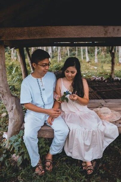 A man and woman sitting together on a wooden bench in a rustic setting, looking at leaves in the woman's hand.