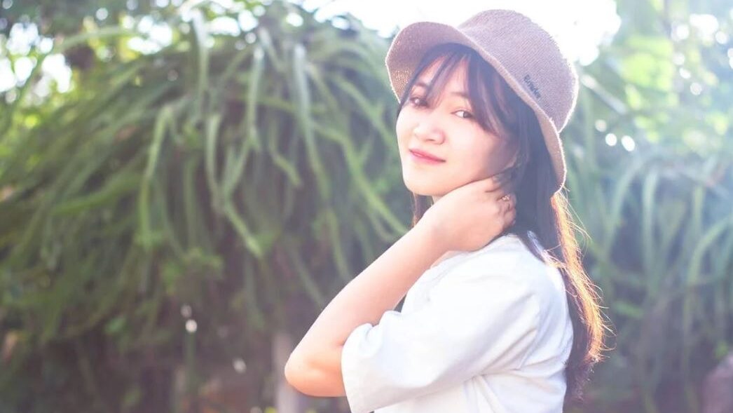 Woman smiling softly while looking at the camera, wearing a hat and basking in the sunlight with greenery in the background.