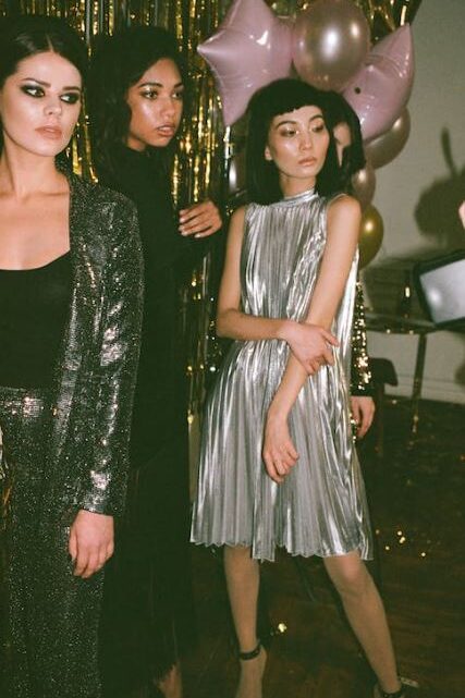 Three women in glamorous outfits, one in a metallic dress and others in shimmering attire, at a social event with balloons and decorations.