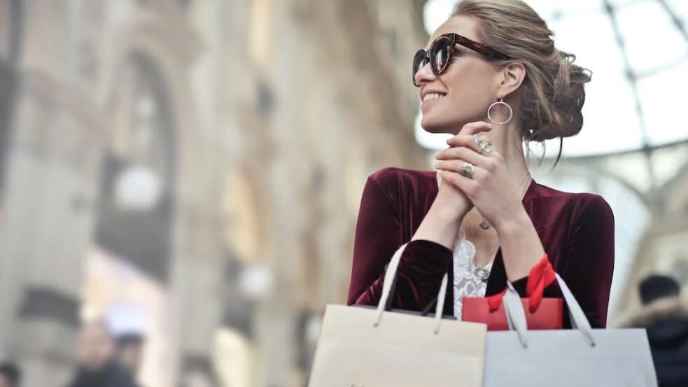 A woman with her hair up and wearing sunglasses smiles while holding shopping bags, enjoying a day out.