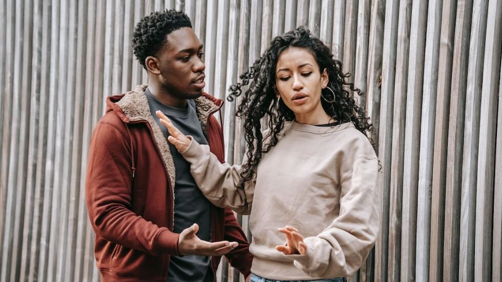 A woman gestures with her hand while a man tries to speak, suggesting a tense conversation by a wooden fence.