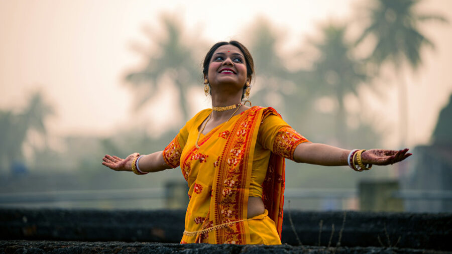 An Hijra in a yellow sari with red embroidery stretches her arms open, smiling against a soft background of trees in the early morning light.