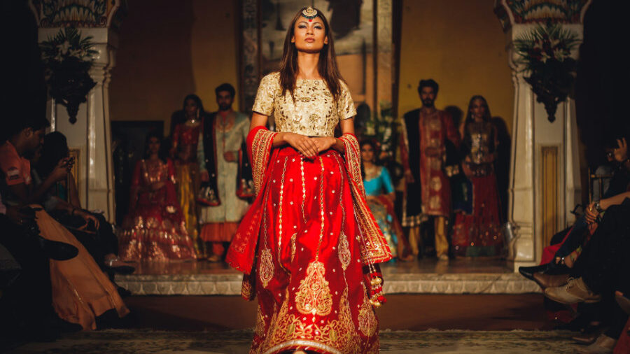 Hijra in elaborate red and gold attire on a fashion runway, with spectators in the background.