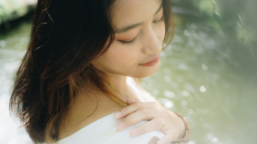 A young Indonesian woman with her eyes closed, gently resting her hand on her shoulder while standing near a natural water feature.