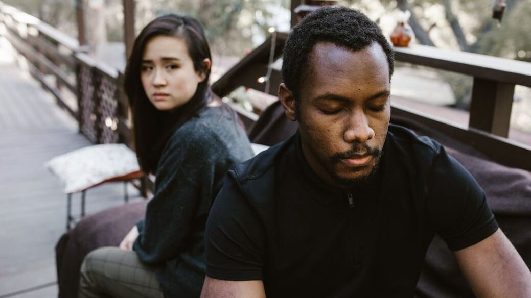 Woman sitting behind a man who looks down, both appearing distant while sitting outdoors on a deck.