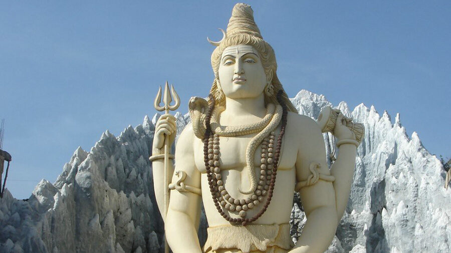 A large statue of Lord Shiva seated in a meditative pose, holding a trident, with a mountainous background.