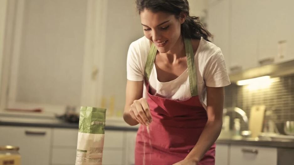 Woman in a home kitchen joyfully adding flour to a recipe, surrounded by ingredients and a warm atmosphere.
