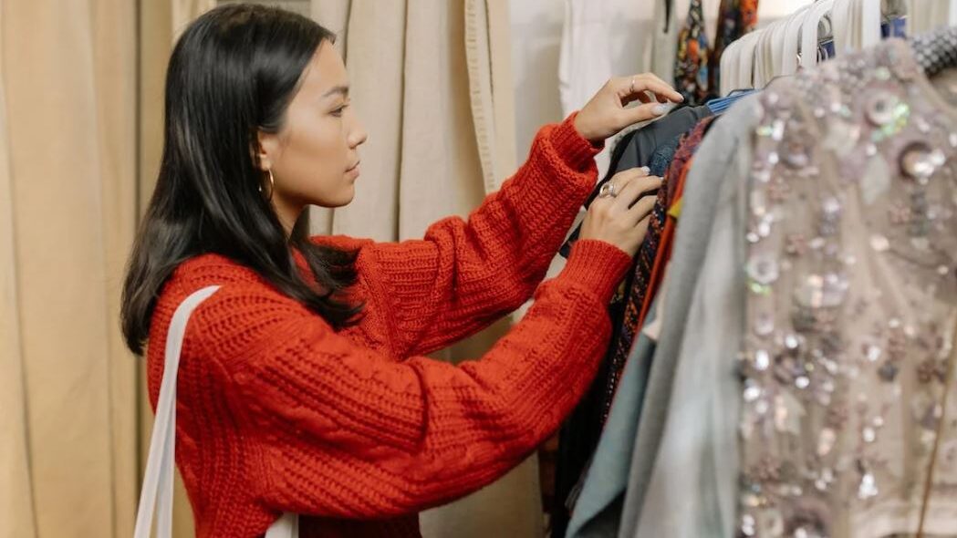 A woman in a red sweater thoughtfully looking through clothes on a rack in a store.