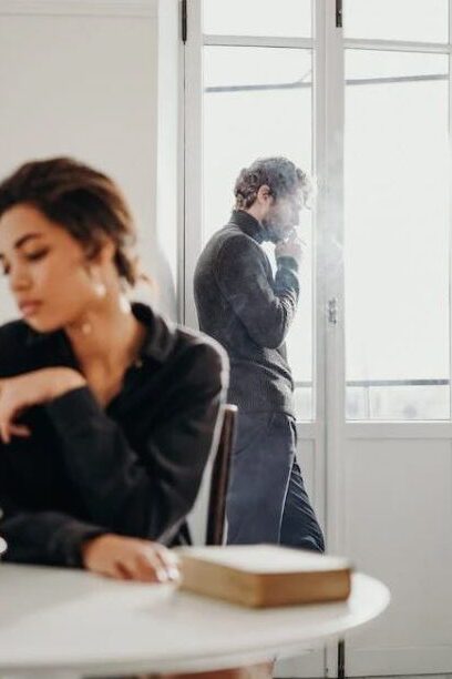 Woman sitting at table with a man smoking near a window, both looking away from each other.