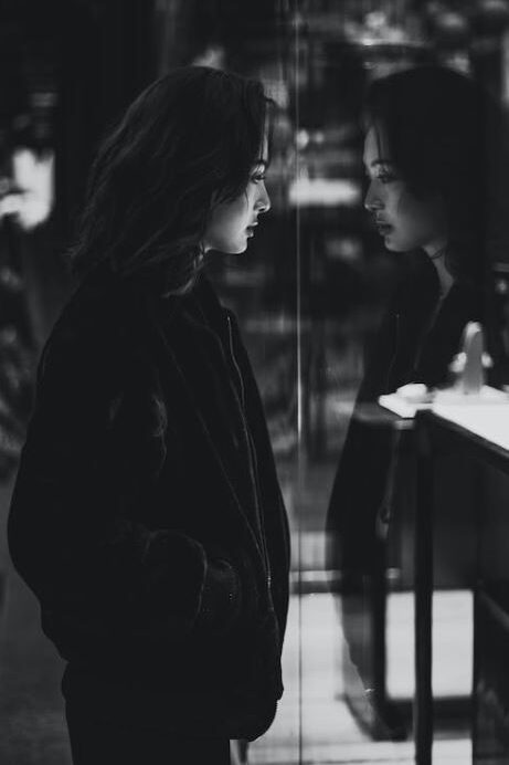 A woman stands in front of a reflective surface, creating a double image effect as she looks at herself in deep thought.