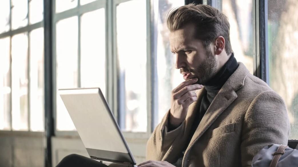 A man in a beige coat and turtleneck, seated by large windows, focused on his laptop with a pensive look and soft natural lighting.