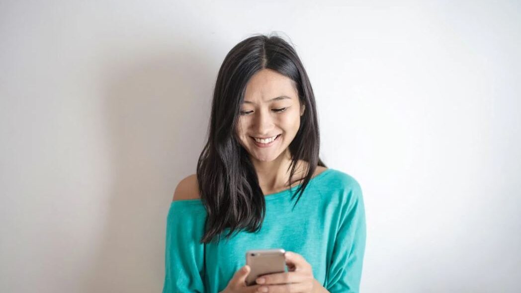 Woman with long dark hair in a teal shirt, looking down at her phone with a happy expression.