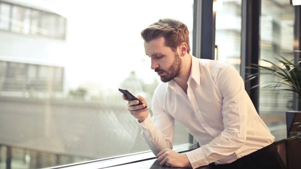 A man in a white shirt, holding his phone and gazing at the screen, with an urban setting visible through the large glass windows.