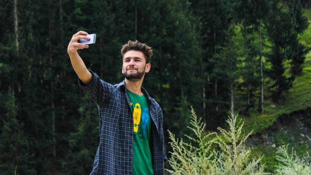 Man taking a selfie in a forested area, wearing a green shirt and plaid overshirt.