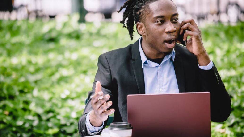 A man in a suit speaking on a phone while gesturing, seated outdoors with a laptop and coffee cup.