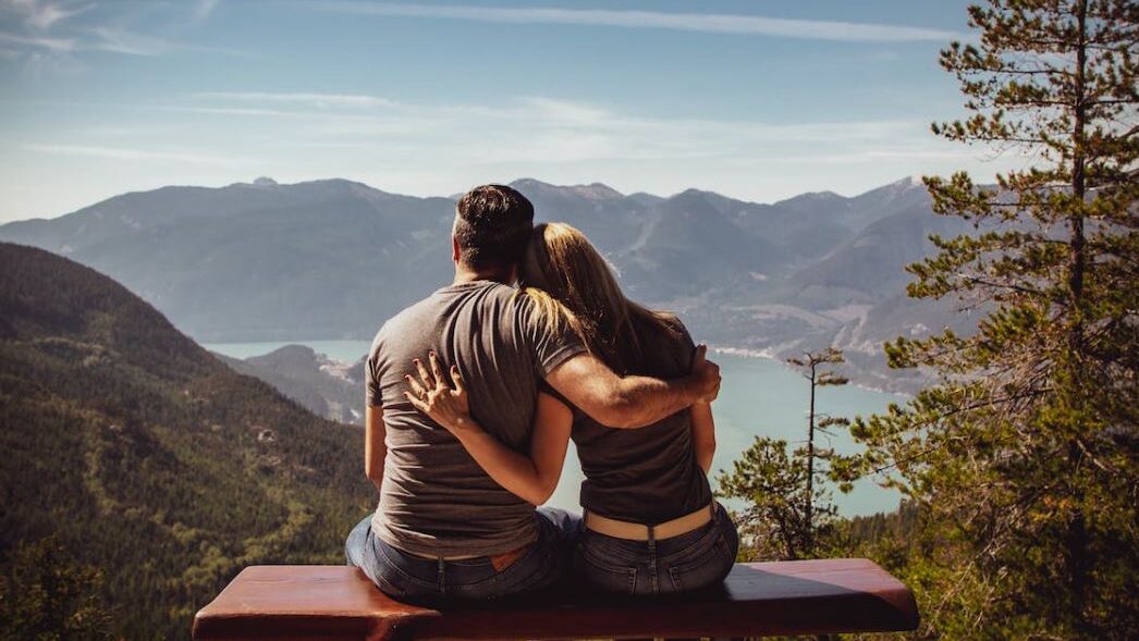 A couple sitting on a bench, embracing, overlooking a scenic mountain and lake view.