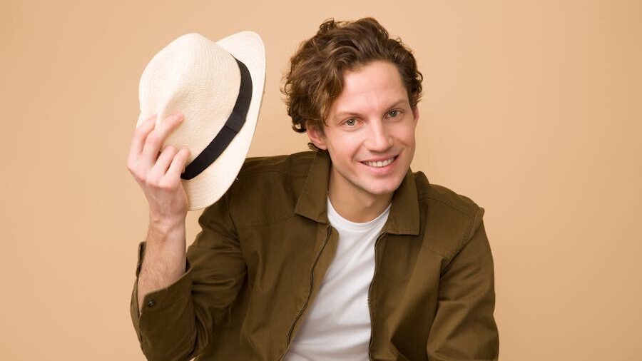 A relaxed and stylish young man wearing a green jacket, playfully posing with a hat in front of a plain beige backdrop.