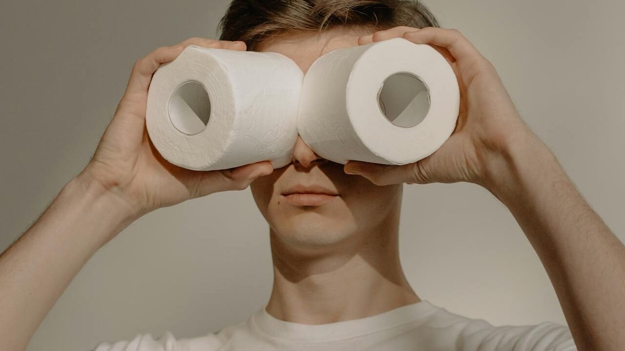 A person holding two rolls of toilet paper up to their eyes like binoculars, with a neutral background.