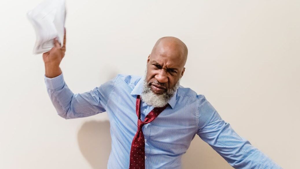 Bald man with a gray beard raising papers in an emotional display against a plain white background.