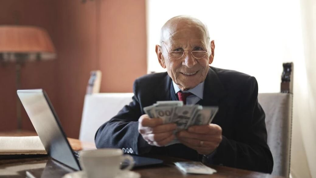 An elderly man in a suit sitting at a table, smiling while holding a stack of hundred-dollar bills, with a laptop and coffee cup nearby.