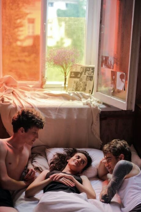 Three young adults relaxing together on a bed in a cozy room with soft lighting, an open window, and warm-colored decor; one person lies in the middle while the others sit on either side, engaged in conversation.