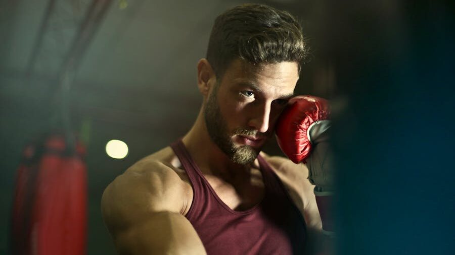 A boxer in red gloves and a matching tank top, staring intensely toward the camera, with blurred gym equipment in the background.