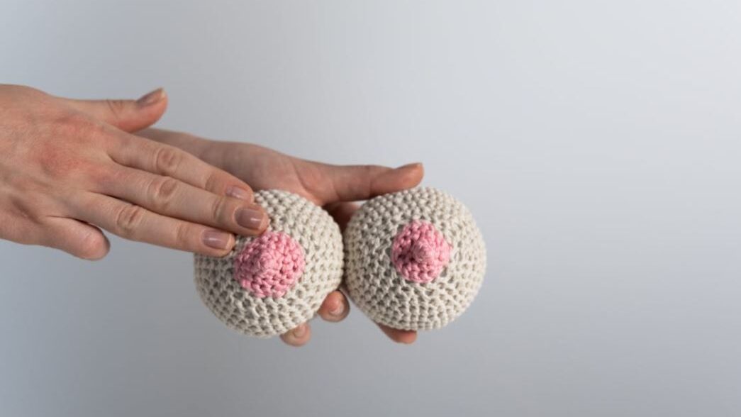 Two hand-made crocheted breast models, one being gently touched by a hand, used for educational or symbolic purposes.